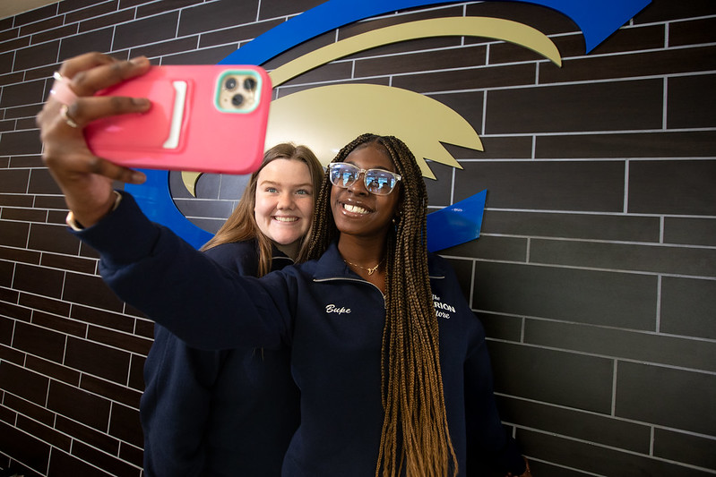 Two young women take a selfie in front of the Golden Eagle emblem in the student center