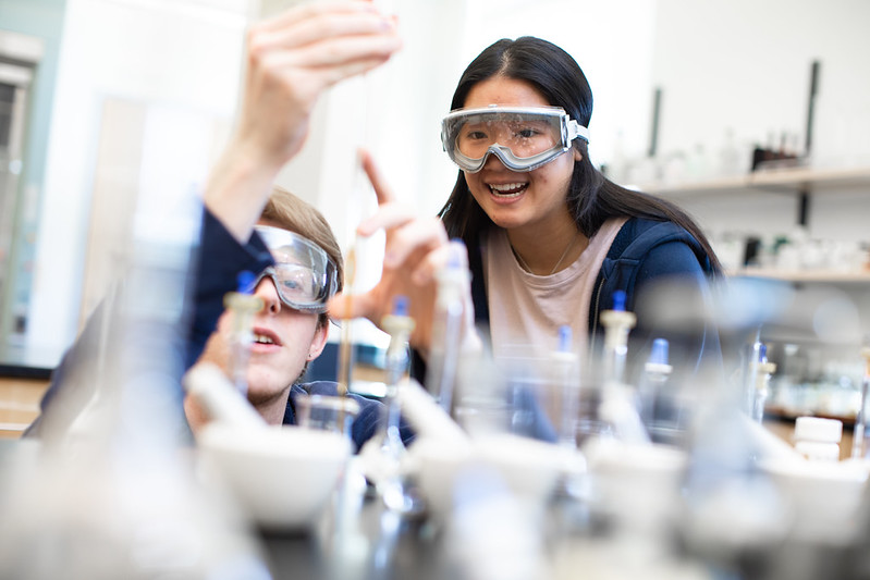 Two students in a chemistry lab wearing goggles conduct an experiment